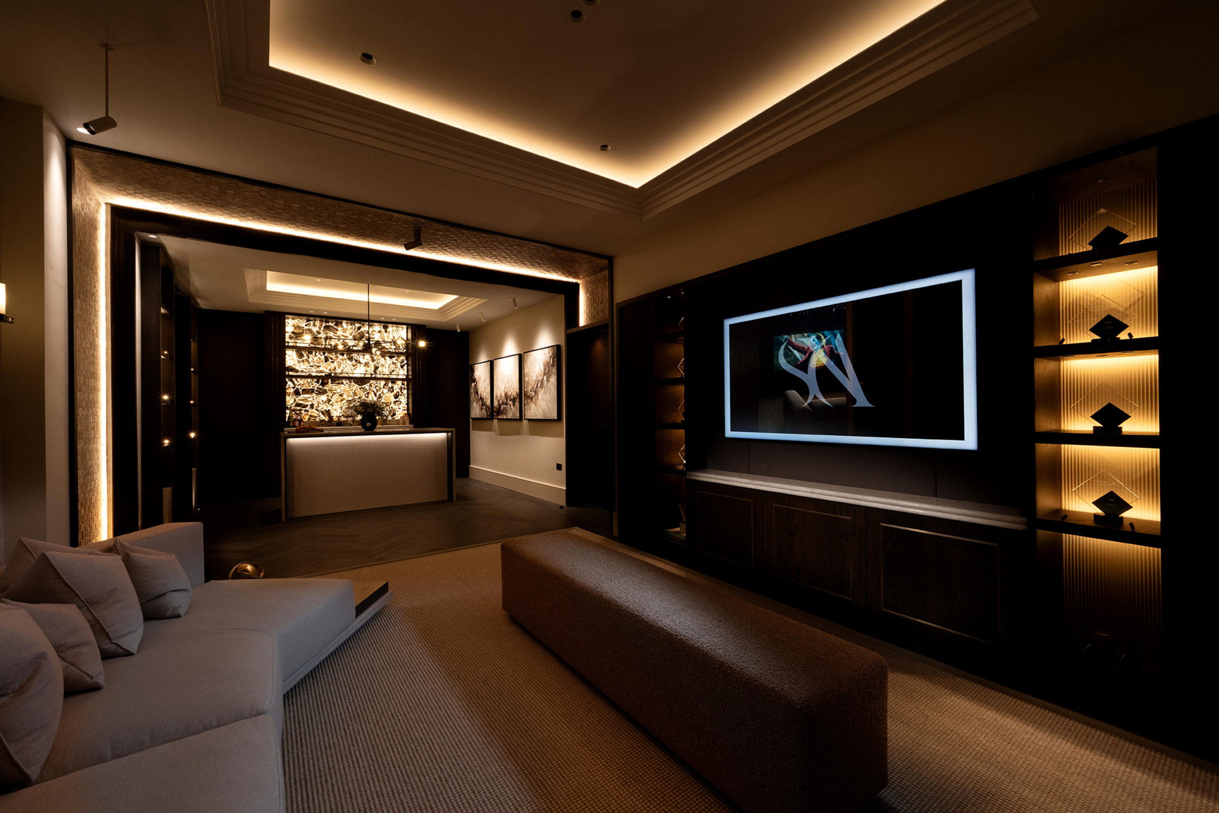 Luxurious dimly lit home cinema lounge with warm recessed cove lighting, a low grey sectional sofa and long ottoman on textured carpet facing a large wall-mounted screen framed by illuminated shelving, and a backlit stone feature wall with a minimalist counter and abstract artwork in the background.