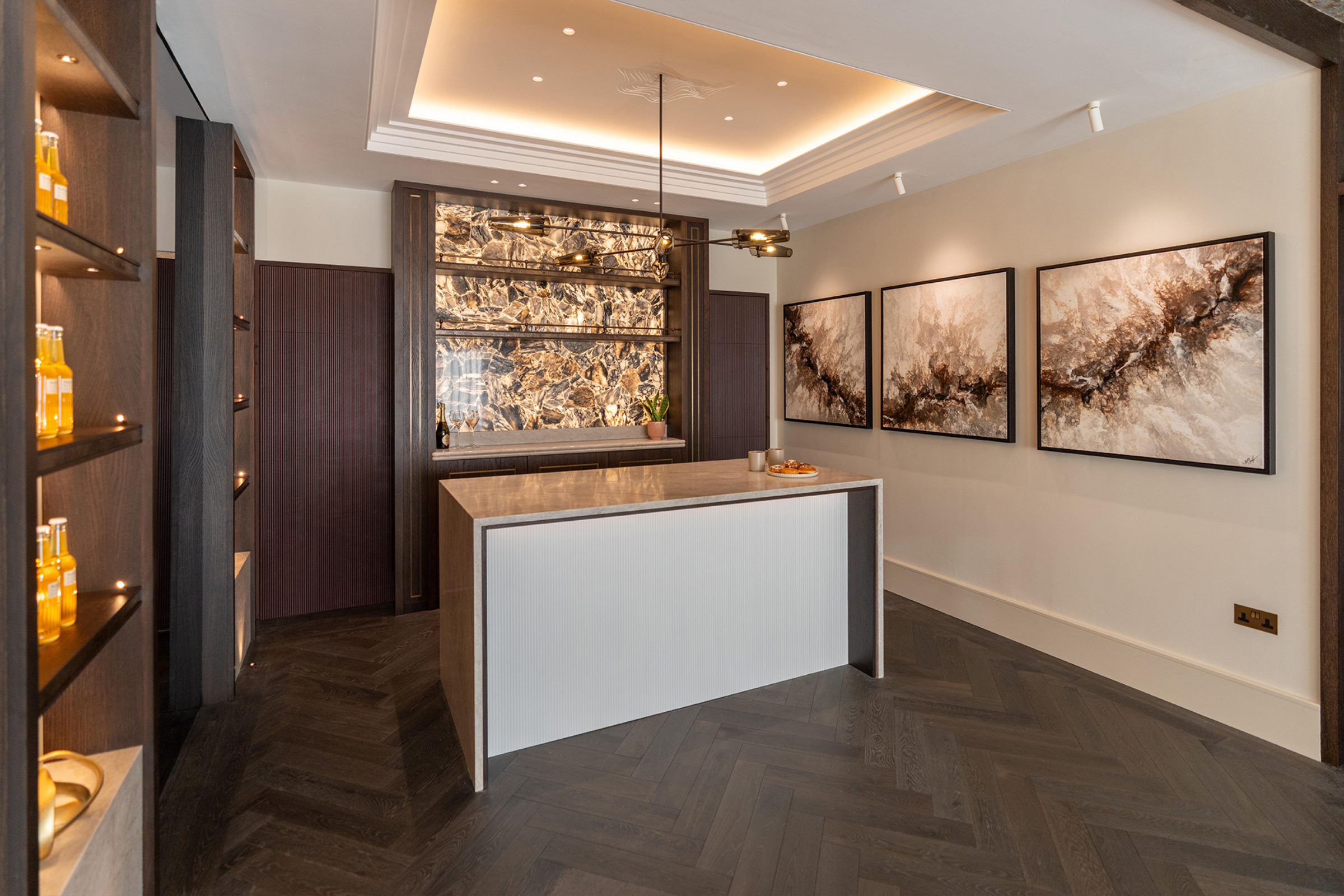 Modern reception area with a central marble‑topped island, backlit veined marble feature wall framed in dark wood, shelving with bottled products on the left, three abstract brown-and-cream paintings on the right, recessed cove lighting and a pendant light above a herringbone wood floor.