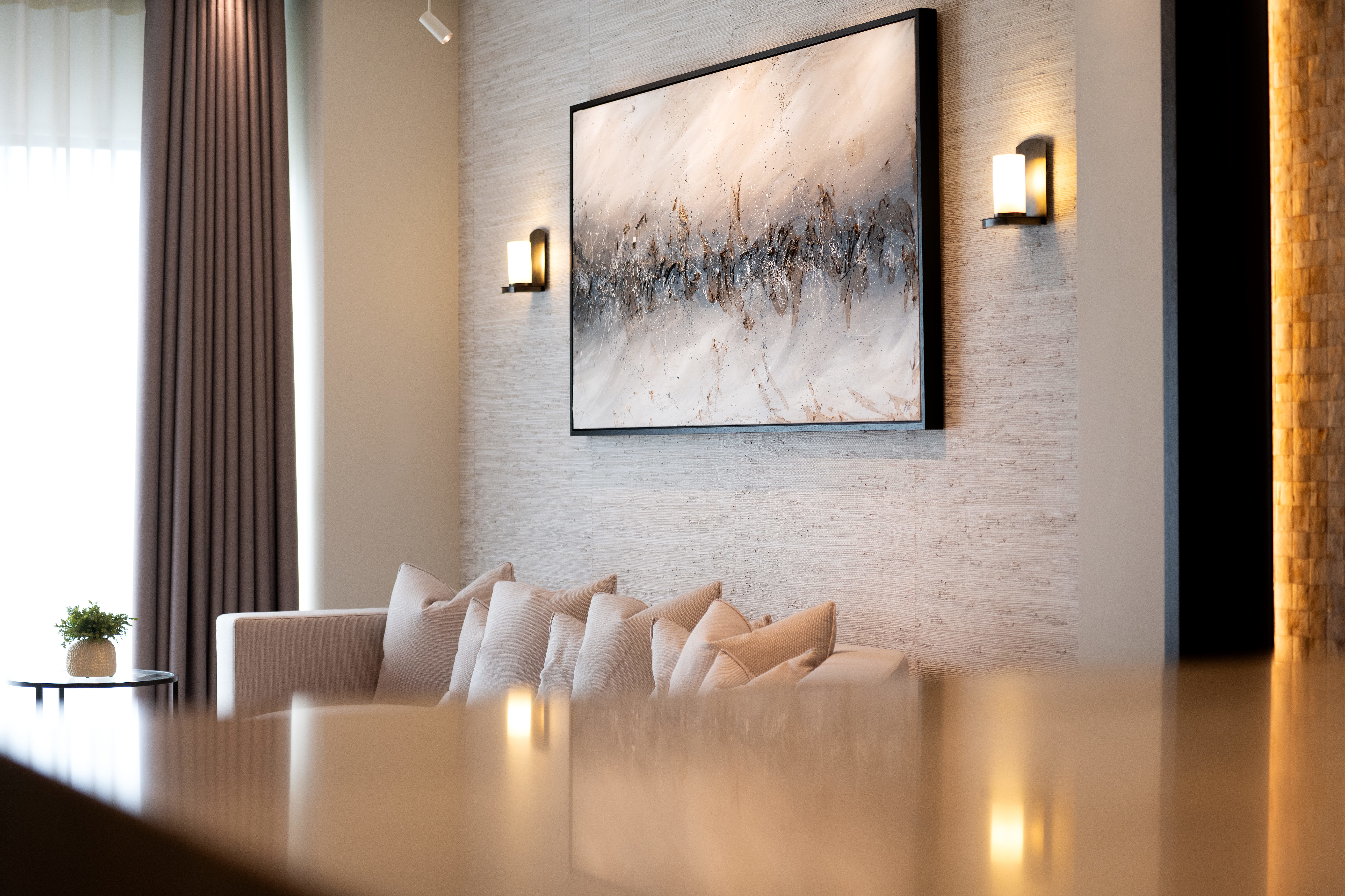 Neutral-toned living room showing a large abstract painting in a thin black frame centered on a textured wall between two lit wall sconces, above a beige sofa piled with cushions, with floor-to-ceiling curtains and a small potted plant on a round side table to the left.