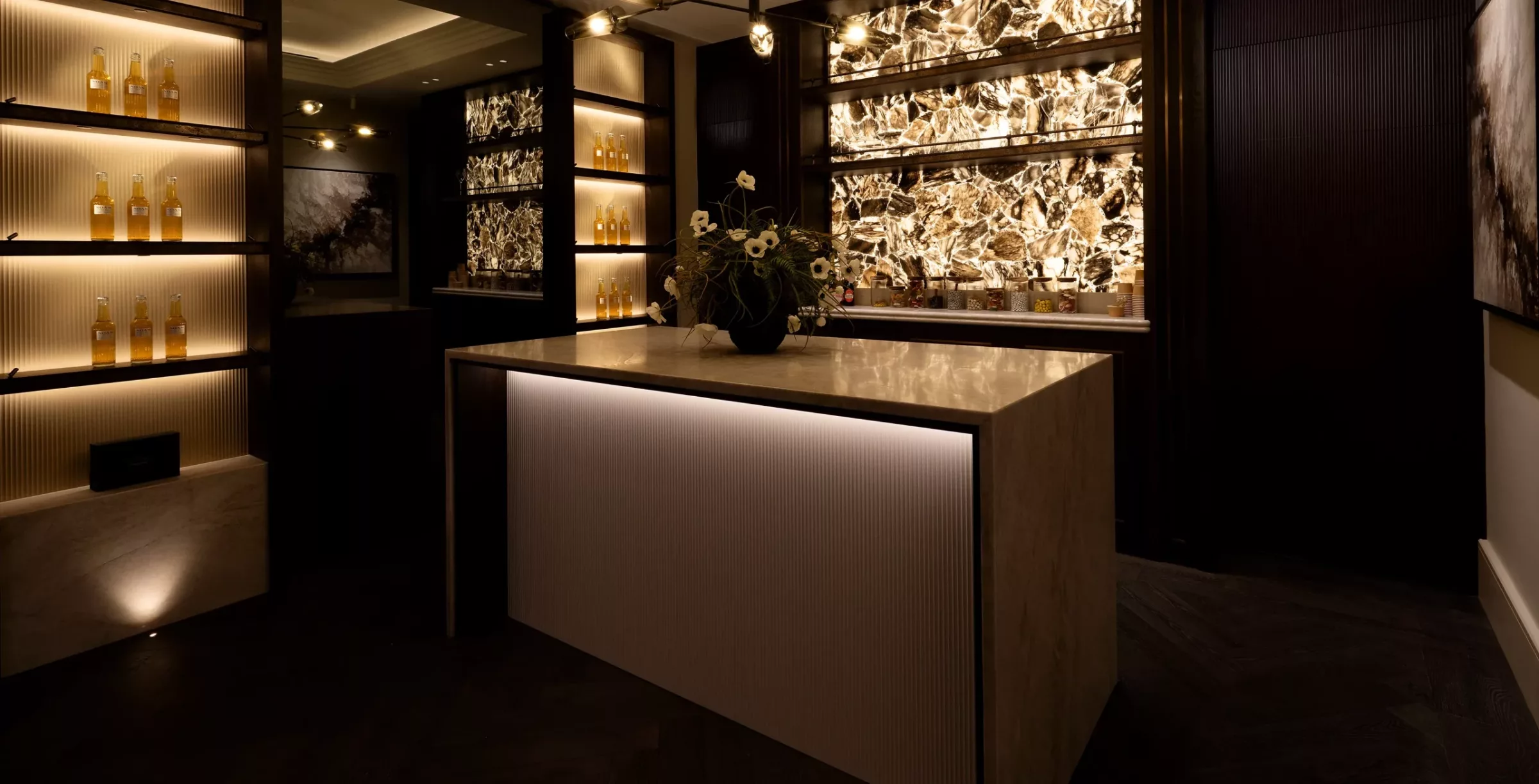 Dimly lit luxury tasting room with a backlit patterned stone wall, a central marble island topped by a vase of white flowers, and illuminated shelving displaying rows of amber bottles and jars of snacks.
